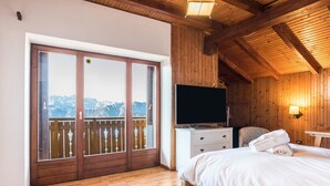 Een slaapkamer met een houten plafond, een bed, een televisie, een dressoir en een balkon met uitzicht op de bergen.