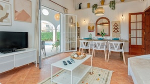 Una sala de estar con televisión, una mesa de comedor con sillas, un sofá y un ventanal corredero que da a un área exterior.