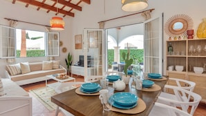 Una sala de estar moderna con una mesa de comedor de madera, sofás blancos y un amplio ventanal con vistas a zonas verdes.