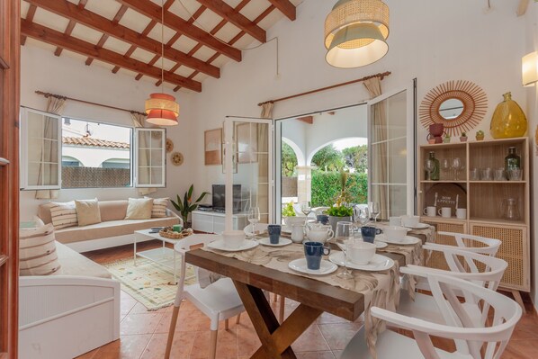 Un coin repas avec une table en bois dressée pour quatre personnes, des chaises blanches et une vue sur le jardin grâce à des portes vitrées.