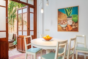 Een eethoek met een ronde tafel, stoelen, een fruitschaal, een schilderij en een houten deur met ruitpanelen.