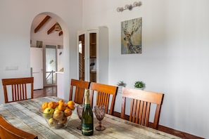 Una mesa de comedor con un frutero, una botella de vino y copas. Alrededor de la mesa hay sillas de madera. Se ve un armario y un cuadro al fondo.