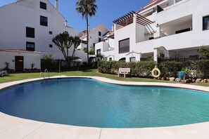 A swimming pool surrounded by white buildings with balconies, a palm tree, and a hedge.