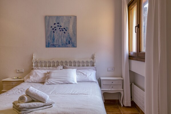 Een slaapkamer met een bed, witte beddengoed, een nachtkastje, een wanddecoratie en een raam met gordijnen.