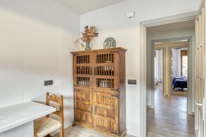 Une pièce avec un meuble en bois, une table blanche et une chaise. On aperçoit un lit à travers une porte ouverte au fond de la pièce.