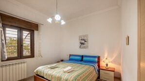 A bedroom with a bed, bedside table, lamp, radiator, window, and artwork on the wall.