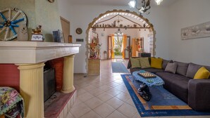 A living room with a sofa, a coffee table, and a fireplace. There is a dining area with a table and chairs. The room has a tiled floor and a large window.