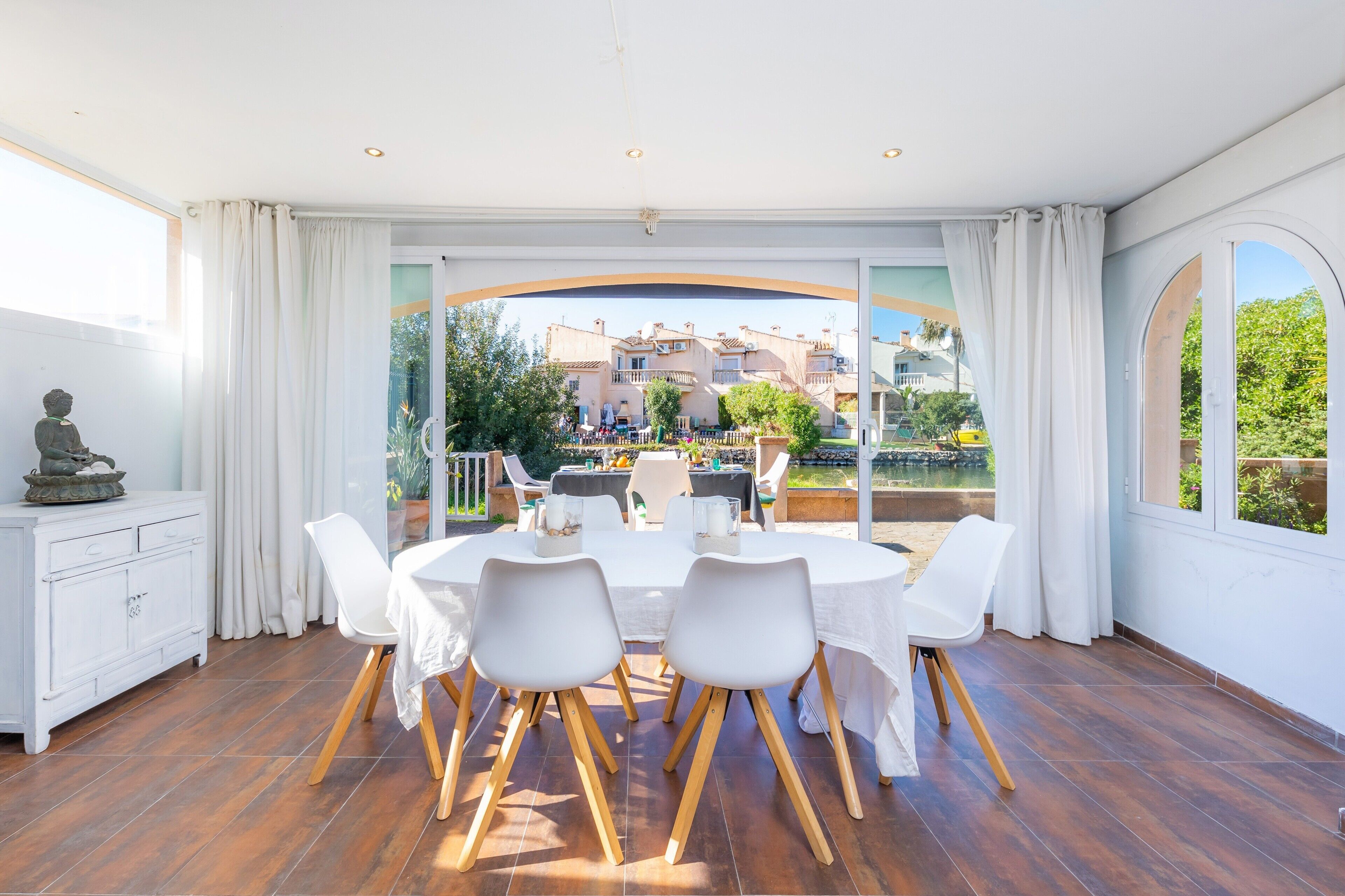 A spacious room with a large dining table, white chairs, and a view of a residential area through large windows.