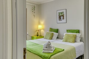 Une chambre d’hôtel avec deux lits, une literie verte, une table de chevet verte avec une lampe, et un tableau encadré au mur.