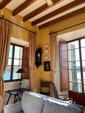 A room with a wooden ceiling, a bed, a table with a lamp, a clock, and a window with curtains.
