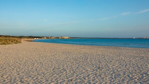 Une plage de sable fin, un ciel bleu dégagé, une mer calme et des bâtiments visibles au loin.