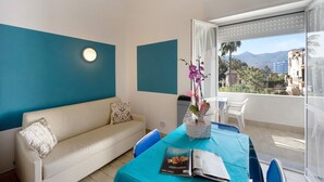 Una habitación con sofá, una mesa de comedor con sillas, un jarrón con flores y vistas al exterior.