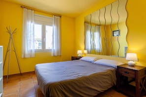 A bedroom with a bed, two bedside tables with lamps, a window with curtains, and a mirror with a wavy design on the wall.