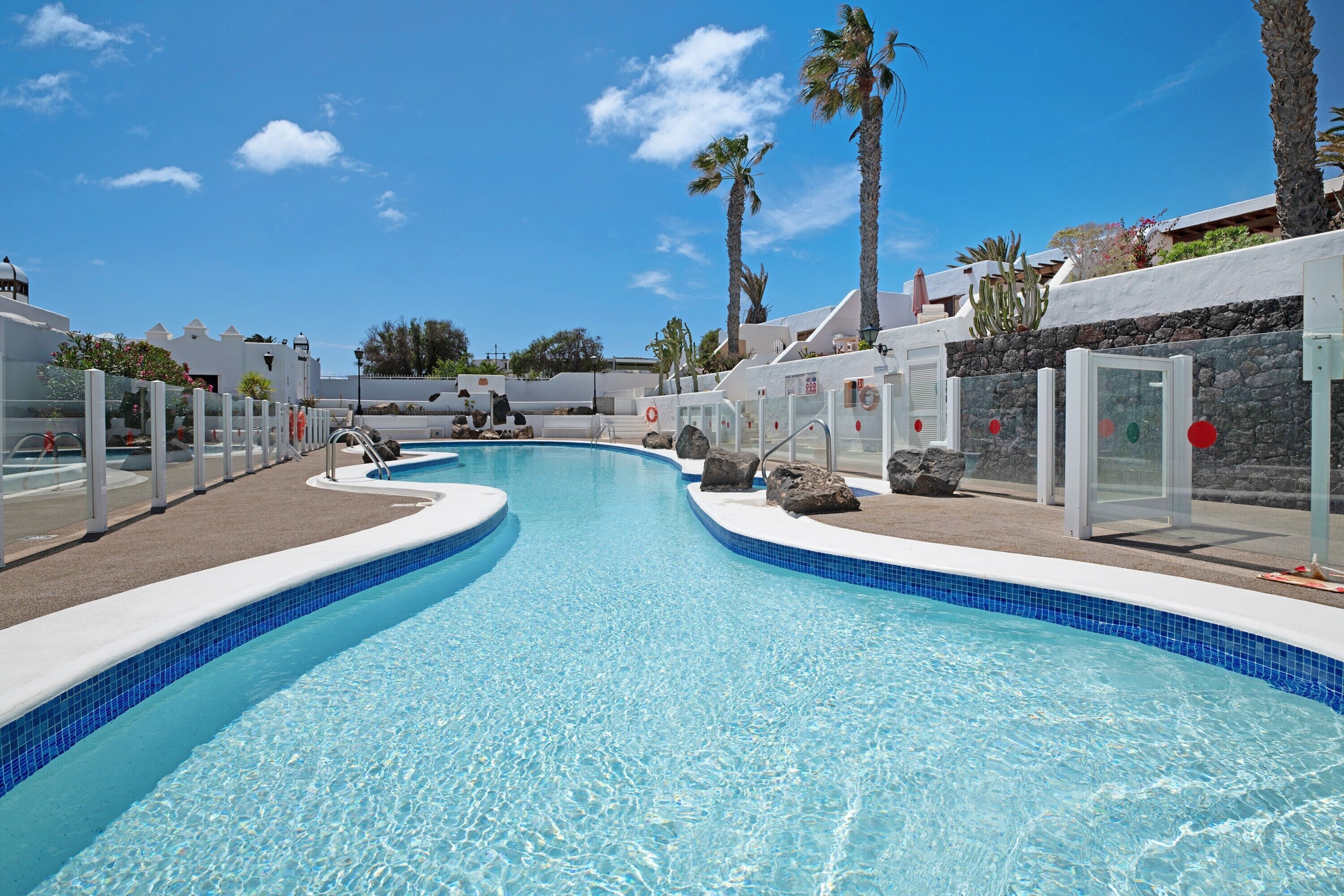 A swimming pool with a curved design, surrounded by palm trees and white buildings.