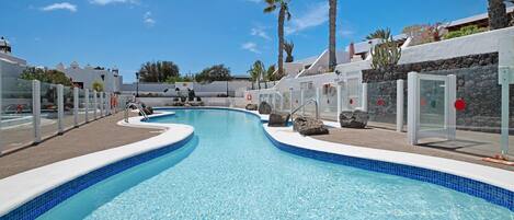 A swimming pool with a curved design, surrounded by palm trees and white buildings.