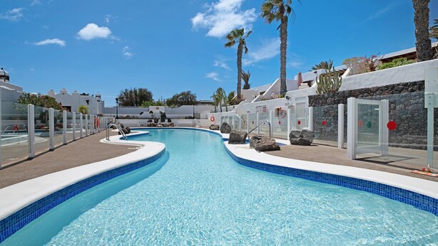 A swimming pool with a curved design, surrounded by palm trees and white buildings.