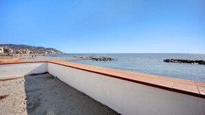 A balcony with a view of the sea, a sandy area, and a white wall with a red border.