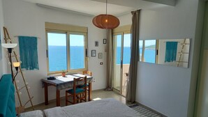 Une chambre avec un lit, une table et des chaises, une vue sur la mer par la fenêtre et un balcon avec un fauteuil.