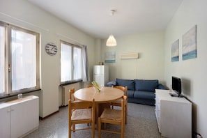 A compact living space with a round dining table, two chairs, a sofa, a TV on a cabinet, and artwork on the walls.