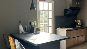 Eine moderne Küche mit einer zentralen Kochinsel, Barhocker, Pendelleuchten und Blick ins Freie durch ein großes Fenster.