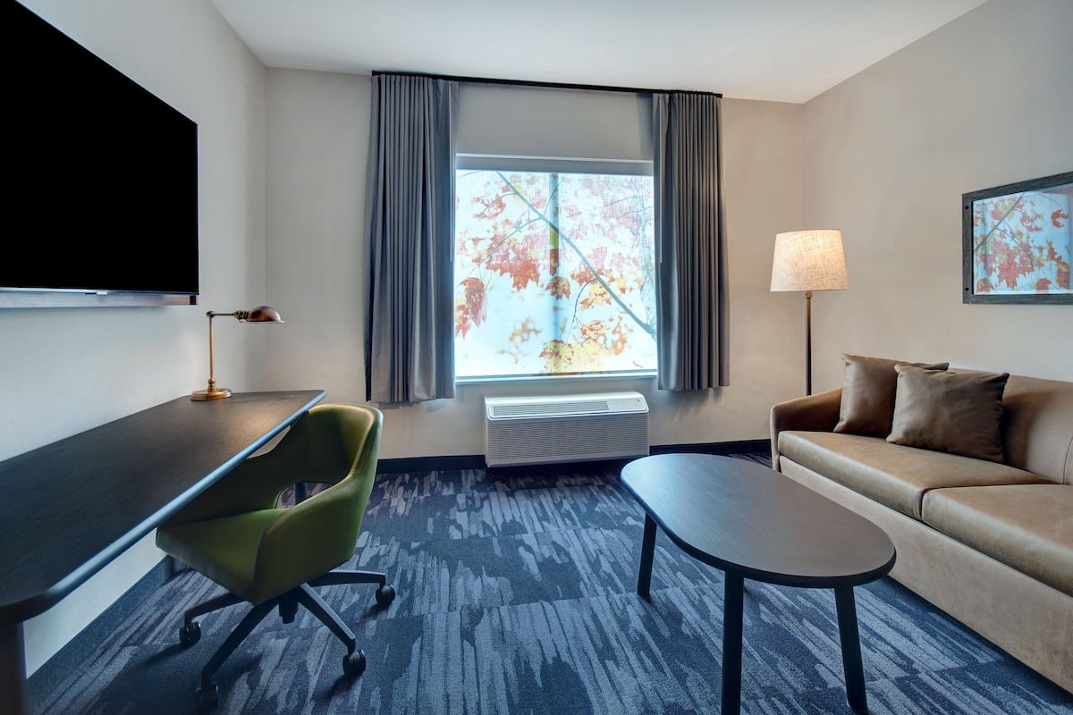 A hotel room with a flat-screen TV, a desk, a green chair, a sofa, a coffee table, a lamp, and a window with curtains.