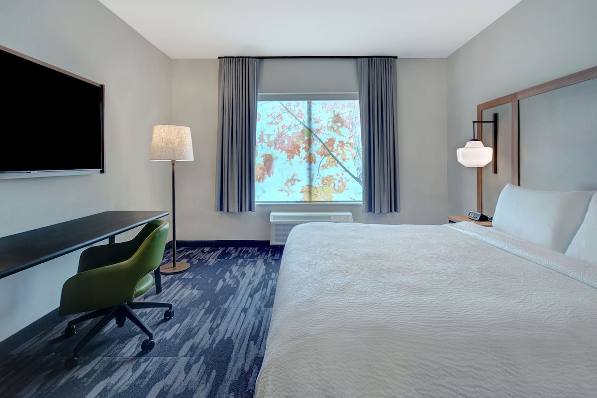 A hotel room with a large bed, a desk with a chair, a flat-screen TV, a lamp, and a window with curtains.