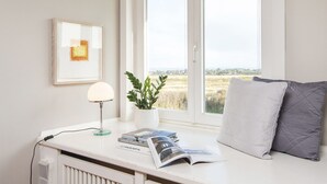 Een kamer met een witte radiator, een lijst met een foto aan de muur, een tafel met een lamp, een plant en een raam met uitzicht op velden.