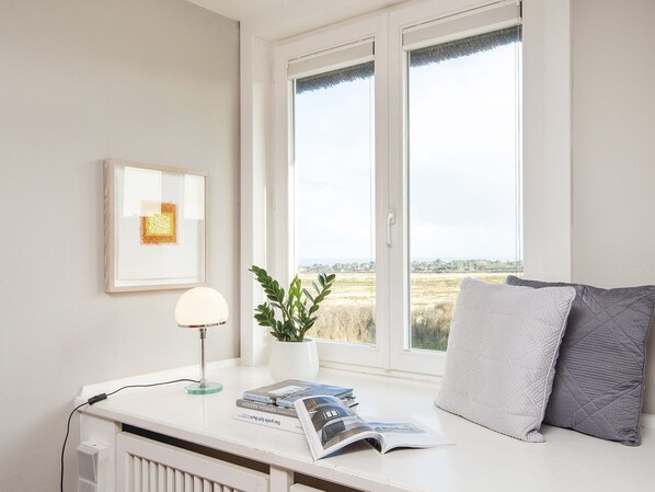 Une pièce avec un radiateur blanc, un tableau encadré au mur, une table avec une lampe, une plante et une fenêtre donnant sur des champs.