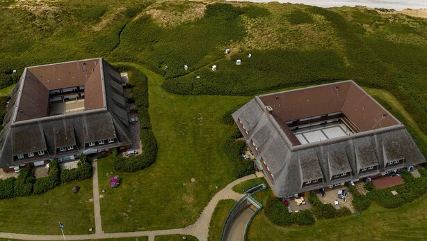 Luchtbeeld van een kustbezit met twee grote gebouwen, een strand en een aangelegde omgeving met paden.
