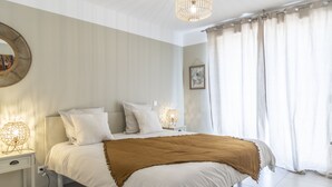 Una cama bien hecha con ropa de cama blanca y una manta marrón, una lámpara de noche y un espejo montado en la pared.