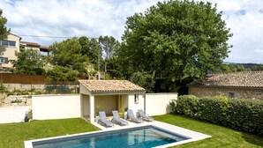 Una piscina rectangular con borde de baldosas, rodeada por un césped cuidado y una pequeña zona cubierta con dos sillones de descanso.