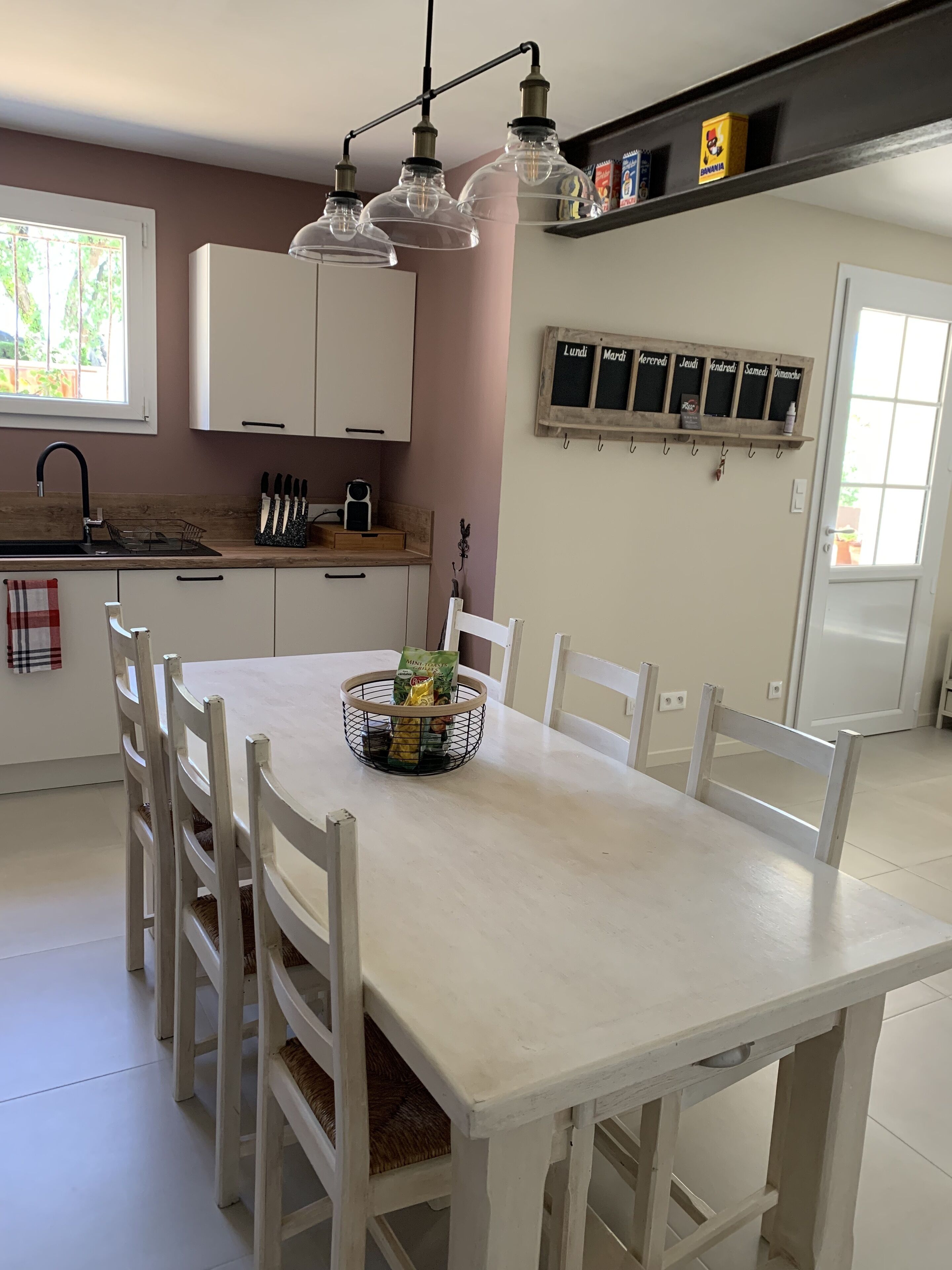 Een keuken met witte kasten, een eettafel met stoelen, een hanglamp en een raam met uitzicht op groen.