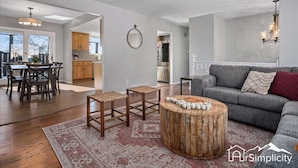 Ein geräumiges Wohnzimmer mit einer grauen Couch, einem runden Holztisch und einem Essbereich mit Holztisch und Stühlen.