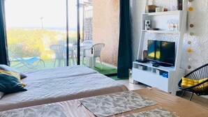 Una habitación de hotel con cama, televisor, estante blanco y balcón con sillas.