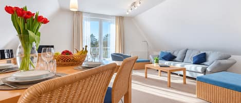 A modern living room with a dining area, featuring a sofa, chairs, a table set for dining, and a view of the outdoors through large windows.