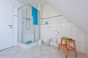 Kylpyhuone, jossa on lasiseinäinen suihkukaappi, wc, puinen jakkara ja pyyheteline.