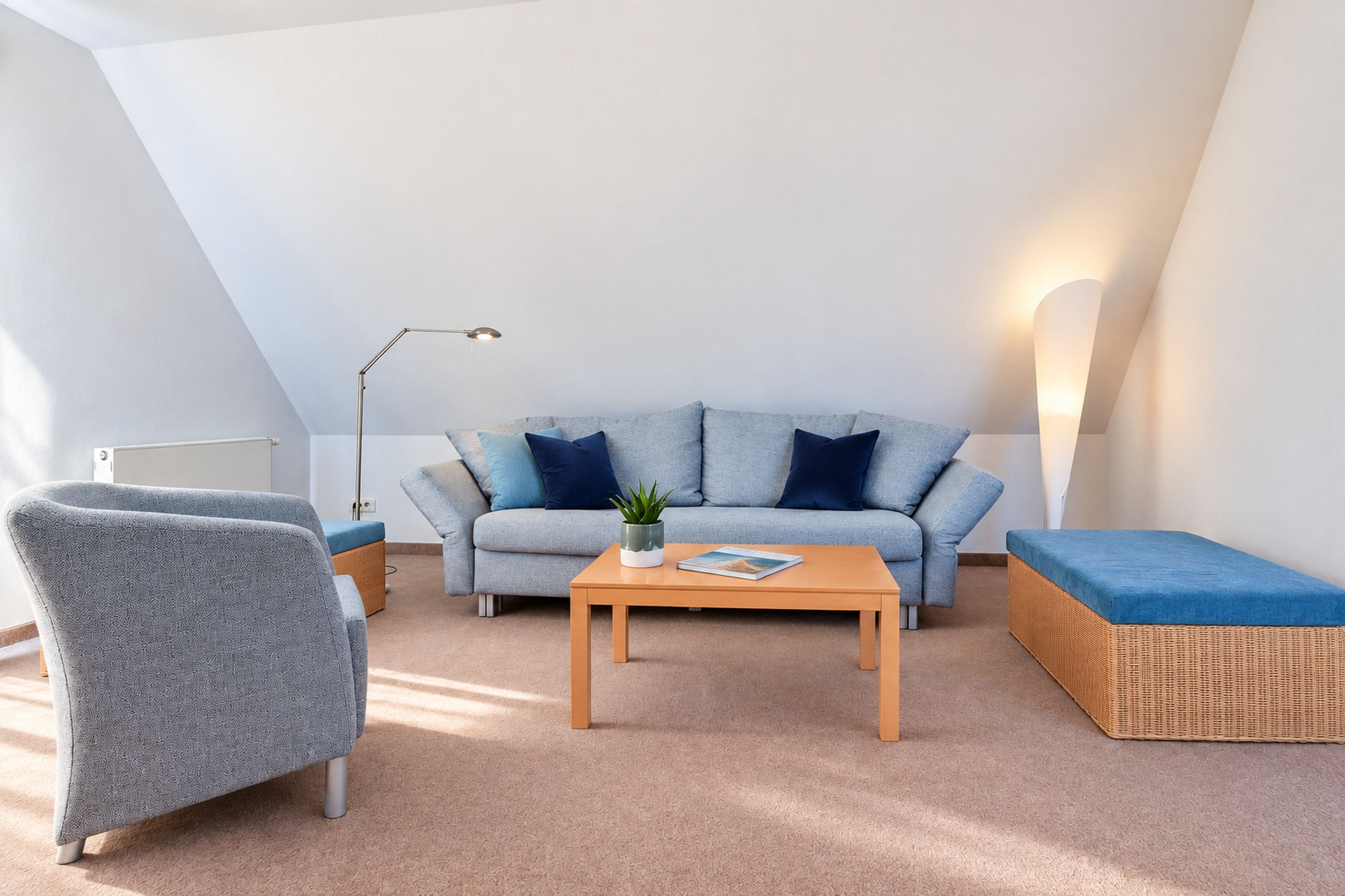 Quarto estilo loft moderno com teto inclinado, um sofá cinza, uma mesa de centro de madeira, uma poltrona cinza e uma mesinha de cabeceira de vime.