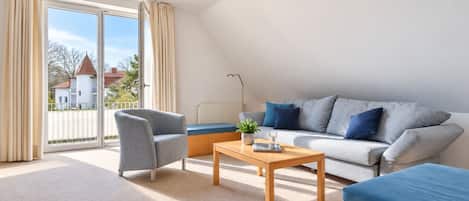Un salon moderne avec un canapé, un fauteuil et une table basse, une grande fenêtre avec des rideaux et une vue sur un quartier résidentiel.