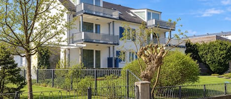 Een meerlaags woongebouw met balkons, een omheinde tuin en volwassen bomen.