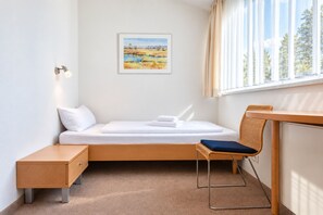 Ein schlicht eingerichteter Raum mit einem Bett, einem kleinen Tisch, einem Stuhl und einem Fenster mit Vorhängen.