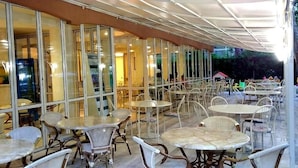 An outdoor cafe with tables and chairs, large glass windows, and a covered area. - Hotel Gioiosa (Cesenatico)