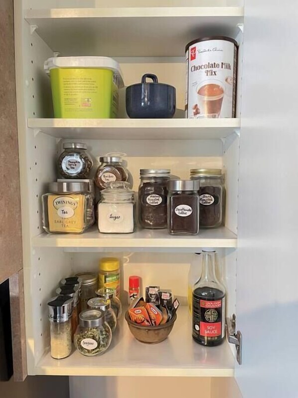 A kitchen pantry with various food items and containers, including jars of spices, a container of chocolate milk mix, and a bowl of cookies.