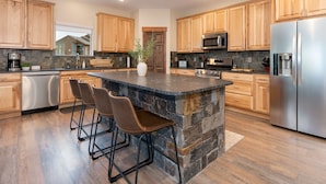 A modern kitchen with wooden cabinets, a large island with bar stools, stainless steel appliances, and a stone accent wall. - Expansive Mountain Views - New Construction, Close to Town + PF, Fireplace, Pool Table, Deck (Grand Lake)