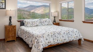 A bedroom with a large bed, two nightstands, and three windows offering mountain views. - Expansive Mountain Views - New Construction, Close to Town + PF, Fireplace, Pool Table, Deck (Grand Lake)