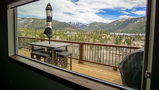 A balcony with a table and chairs, overlooking a mountainous landscape with a lake and buildings. - Expansive Mountain Views - New Construction, Close to Town + PF, Fireplace, Pool Table, Deck (Grand Lake)