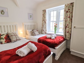 Ein Hotelzimmer mit zwei Betten, jeweils mit roter Decke und weißen Handtüchern, einem Fenster mit floralem Vorhang und einer Vase mit Blumen auf der Fensterbank.