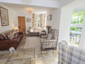 A cozy living room with a brown leather sofa, a grey sofa, and a plaid armchair. There is a stone floor, a rug, and a wooden door.