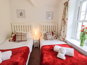 A hotel room with two beds, each with a red bedspread and white pillows. There is a nightstand with a lamp between the beds, and a window with floral curtains.