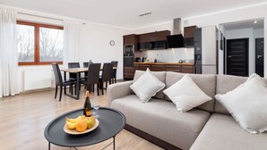 A modern living room with a sofa, a dining table with chairs, a kitchen area with cabinets and appliances, and a large window with curtains.