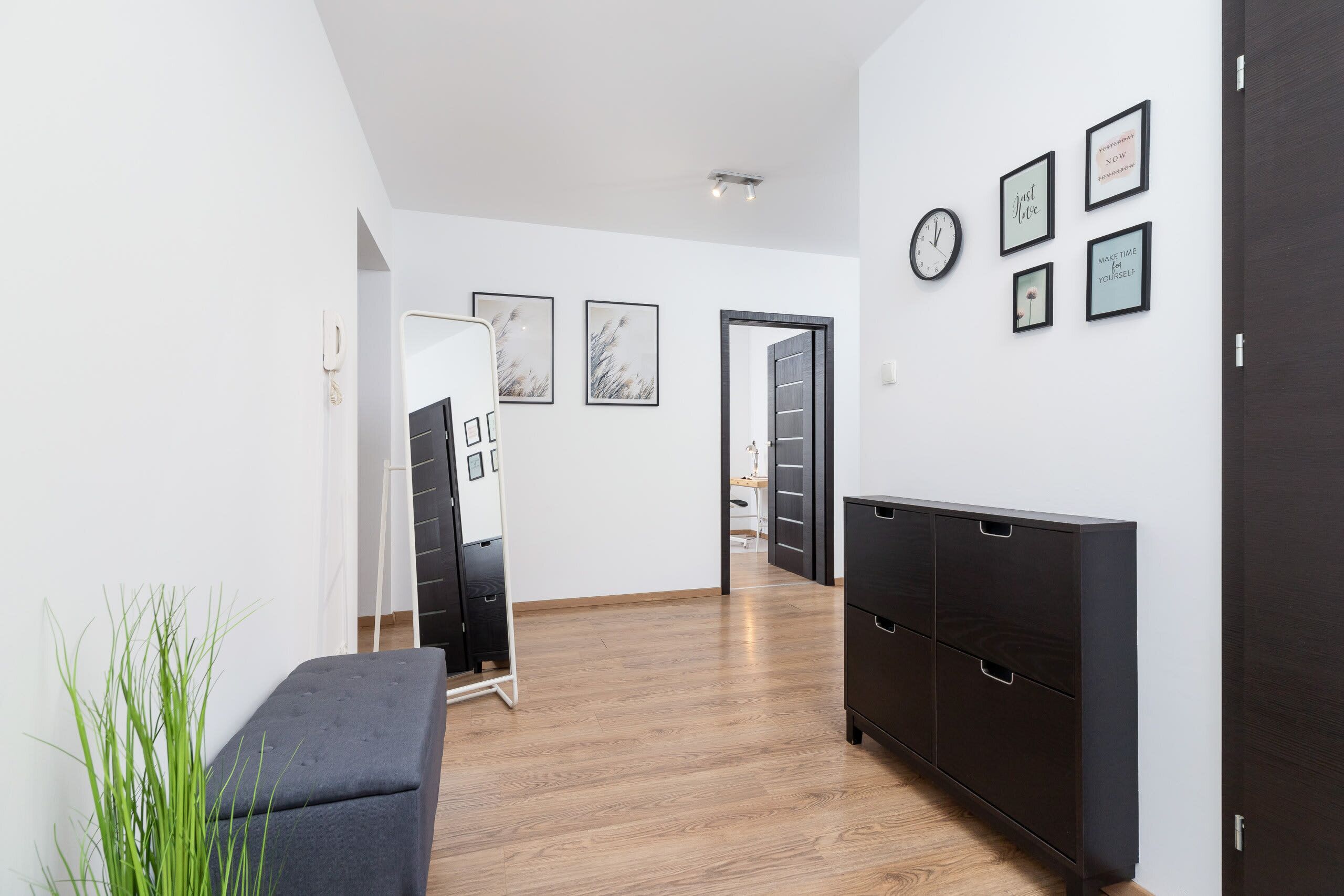 Un interior moderno con suelo de madera, un mueble oscuro y una pared blanca con cuadros enmarcados.
